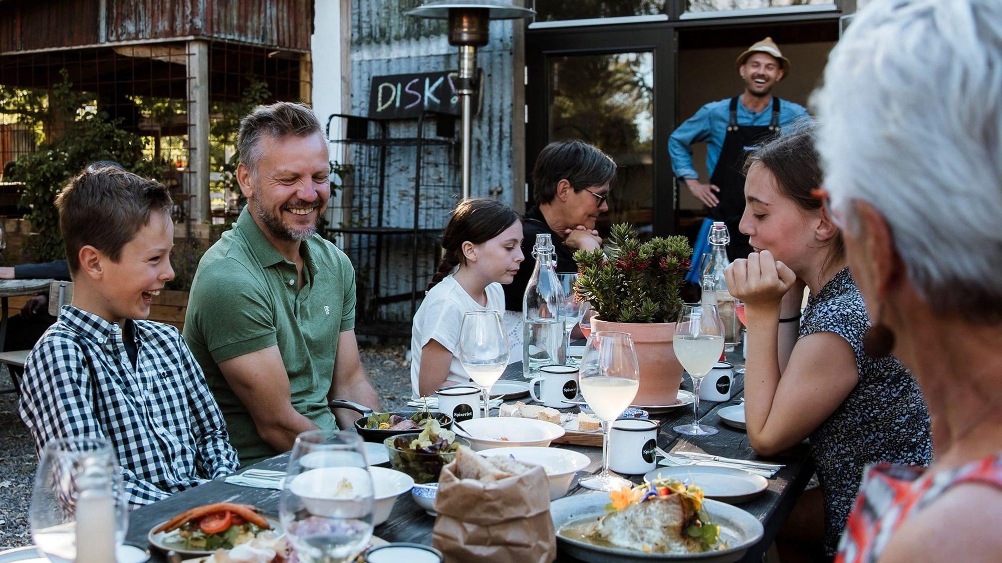 En glad familie sidder på en udendørsservering på restaurant med en glad servitør i baggrunden